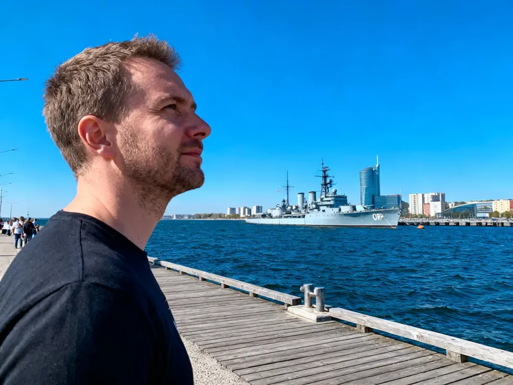 Mężczyzna stojący na drewnianym molo w Gdyni z widokiem na okręt ORP Błyskawica oraz panoramę miasta.