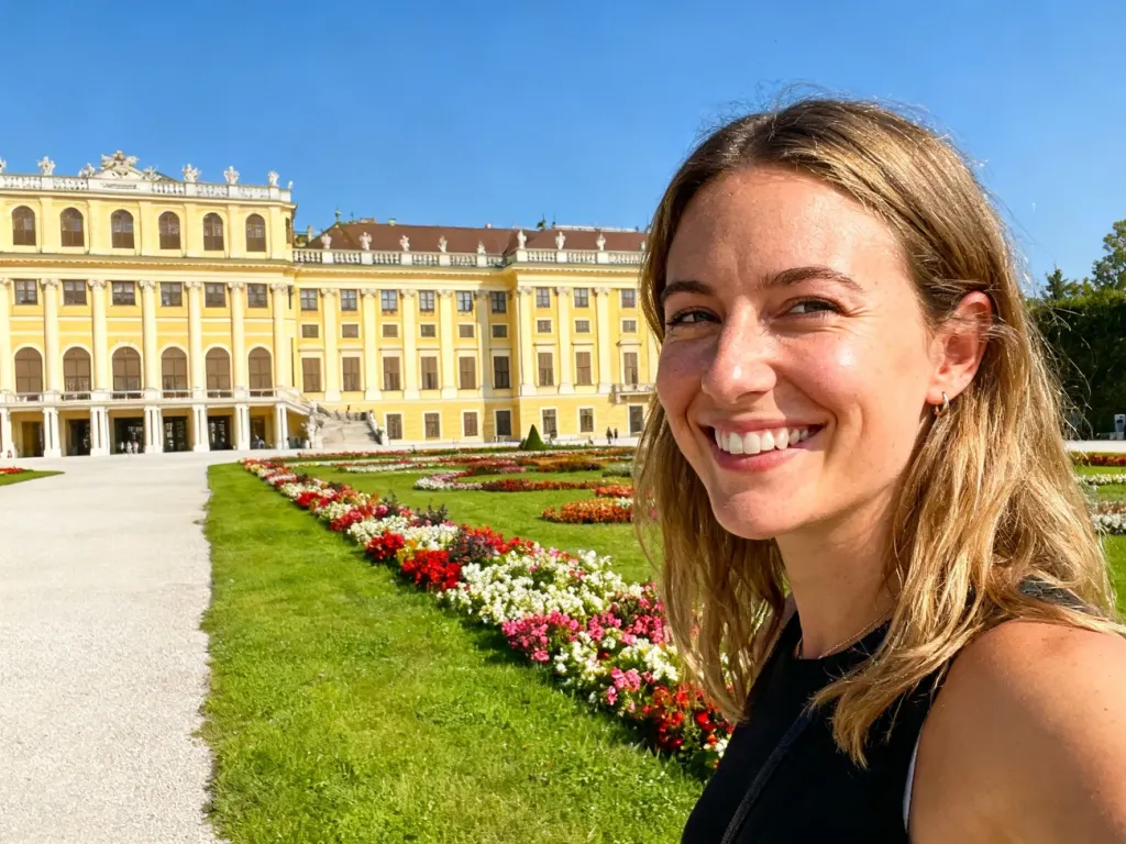 Uśmiechnięta turystka na tle barokowego pałacu Schönbrunn w Wiedniu, otoczonego zadbanymi ogrodami w słoneczny dzień.